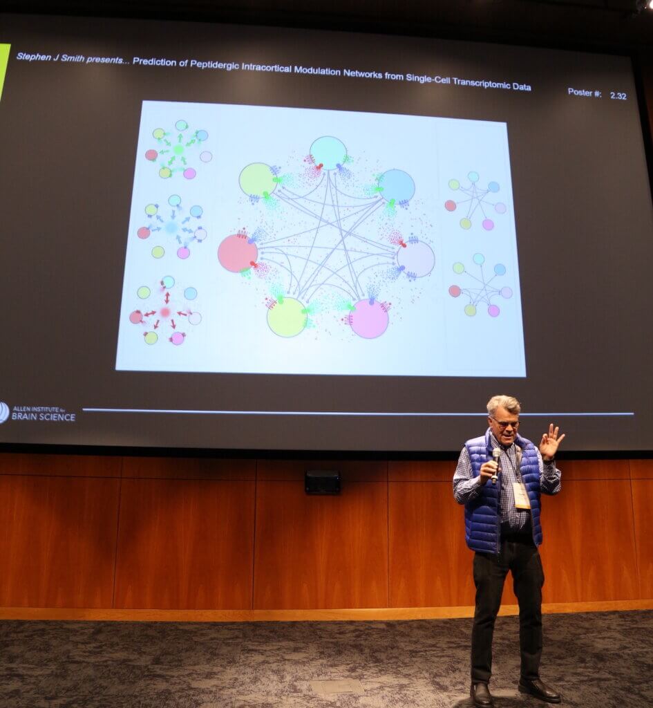 A scientist speaking into a microphone stands in front of a presentation of a diagram of colorful circles with lines connecting them.