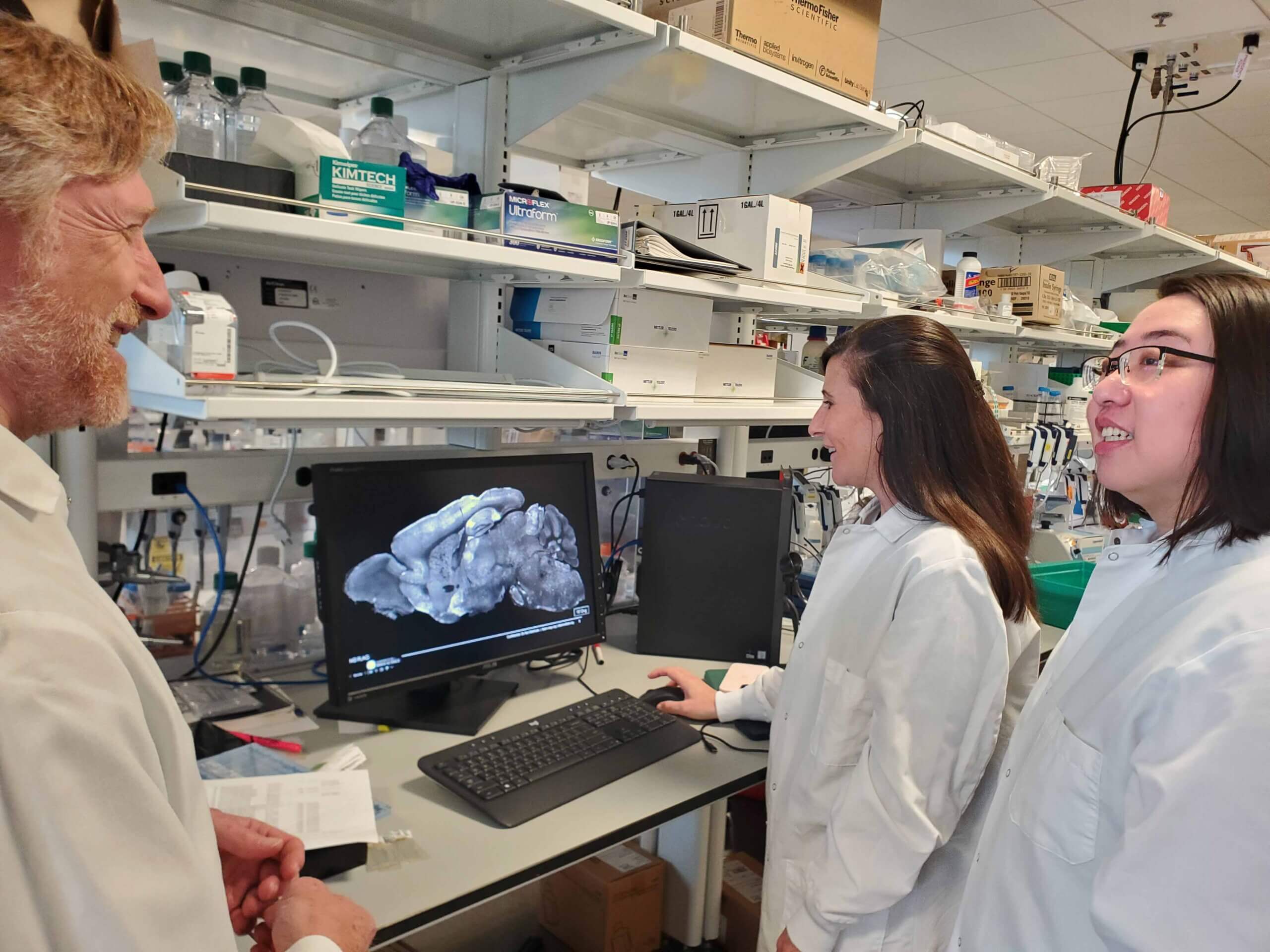 Three researchers in white lab coats talking in front of a computer screen in a lab