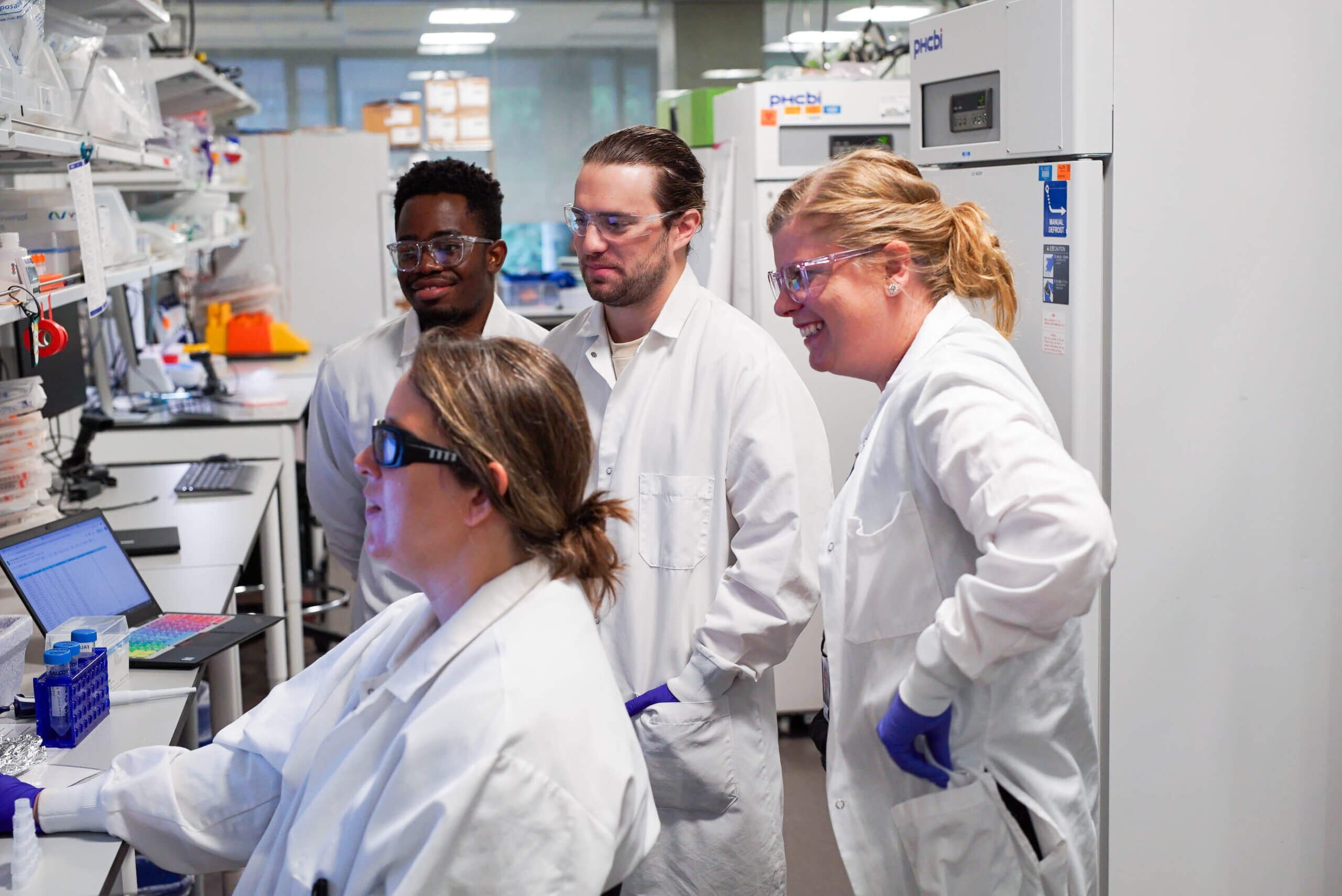 Four researchers wearing white lab coats standing at a table looking at data in a laboratory