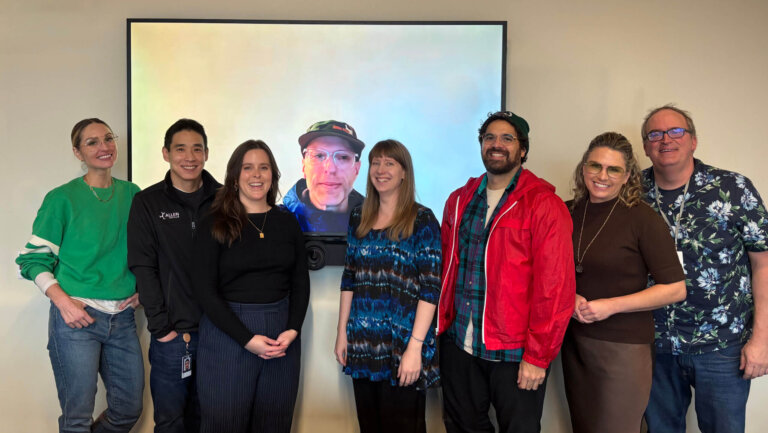 Seven people standing against a wall smiling. Behind them is a large TV monitor with a face staring in